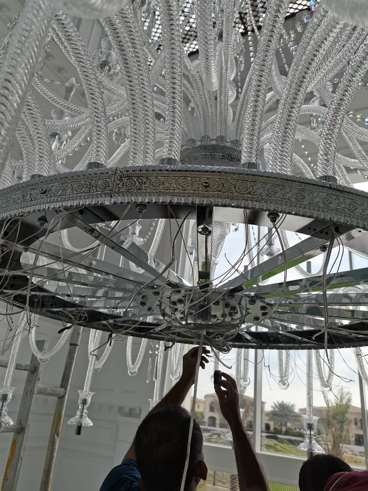 Person assembling a large, intricate chandelier with glass tubes hanging in a bright indoor setting.